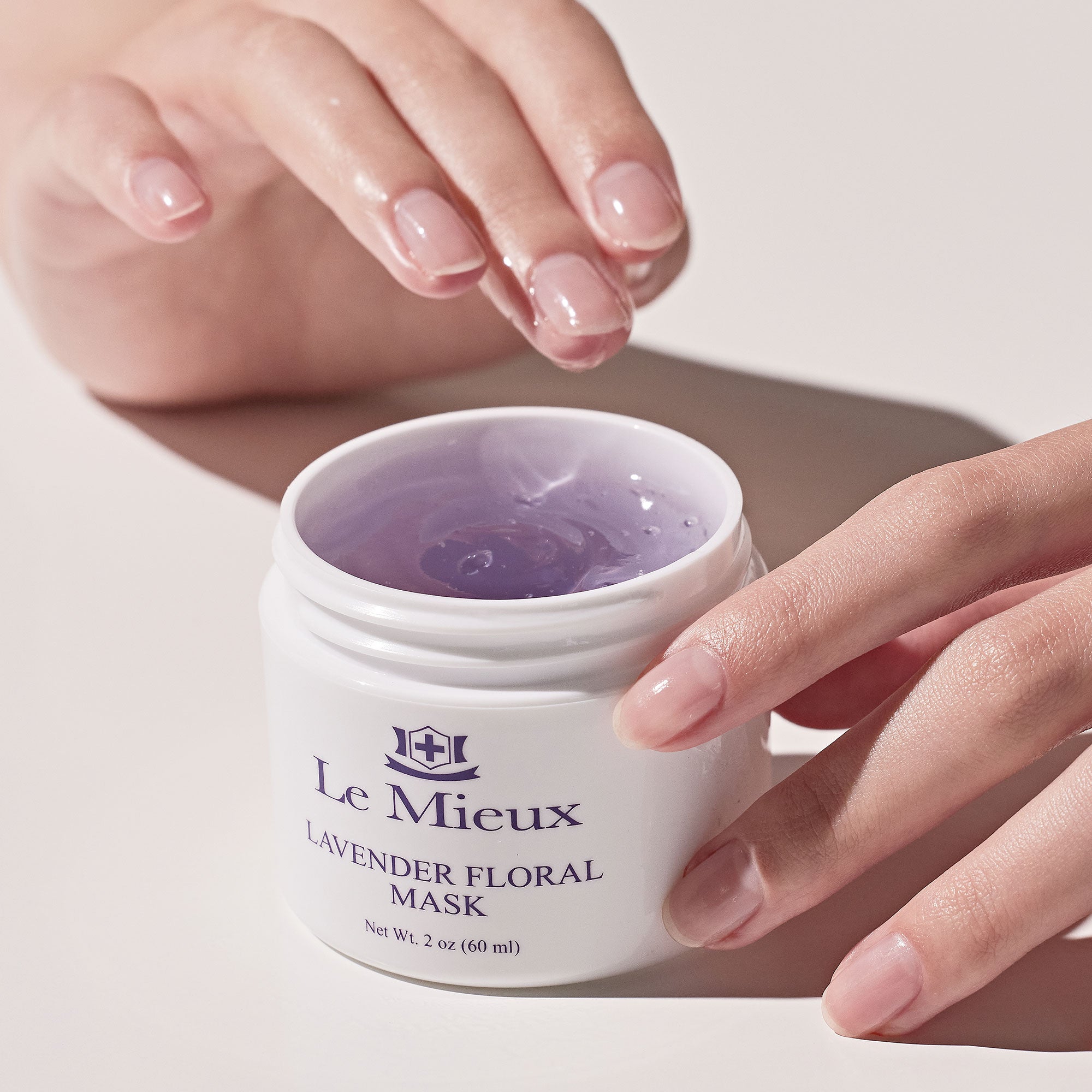 One hand resting against the side of a white lidless container that reads "Lavender Floral Mask" with Le Mieux logo and another hand hovering over it, finger dipped in the light purple gel mask product.
