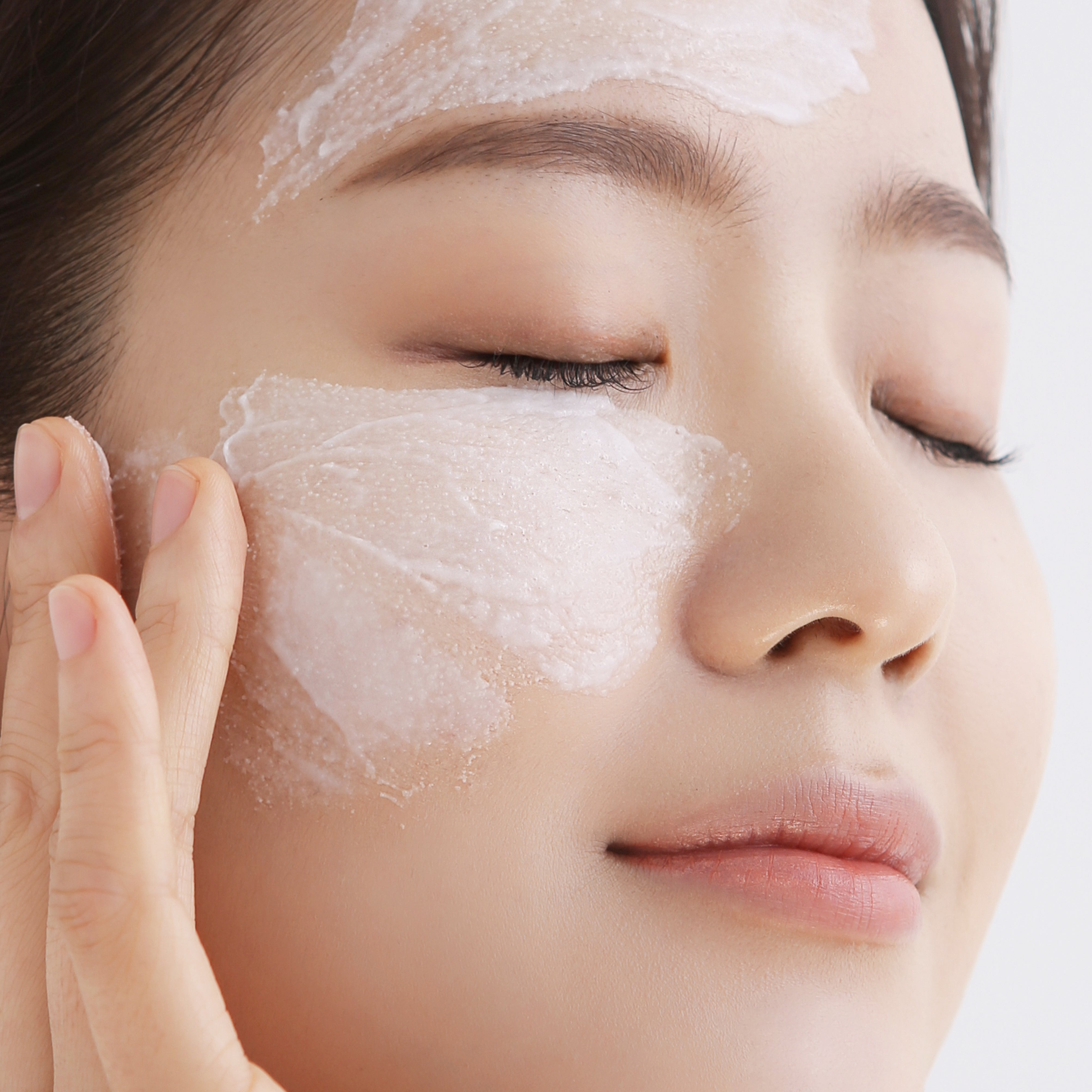 woman with eyes closed, fingers spreading white exfoliant texture onto her cheek and forehead.