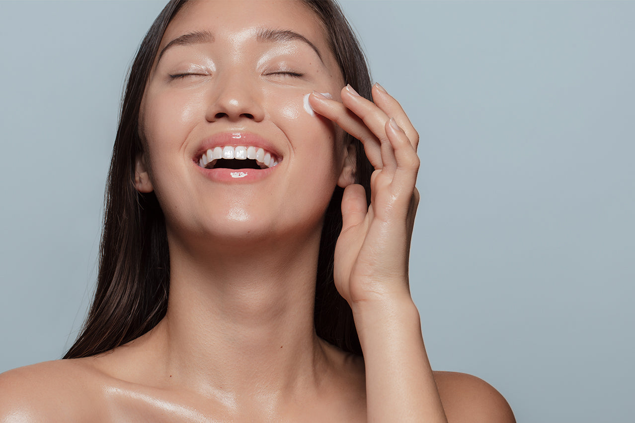 smiling woman with eyes closed, applying white cream to cheek in front of a blue background