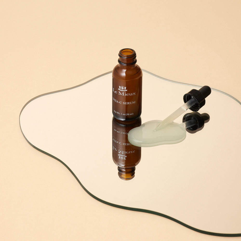 Vita-C Serum next to dropper lid and a pool of serum reflected on a mirror on a light peach background