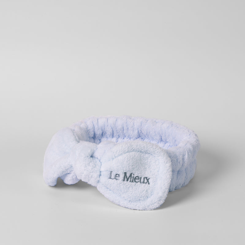 Blue headband with the "Le Mieux" logo on one side of the bow isolated in front of a light gray background.