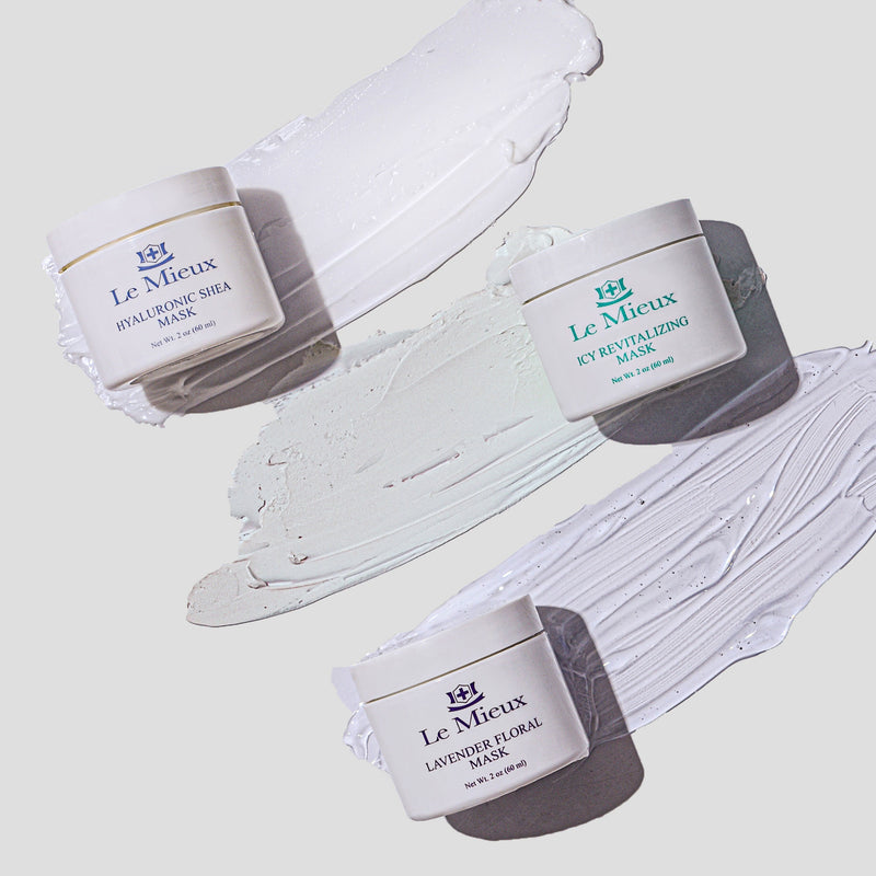 White container that reads "Hyaluronic Shea Mask" with the Le Mieux logo resting on the left end of a creamy texture swab. Underneath is a white container that reads "Icy Revitalizing Mask" with the Le Mieux logo resting on the right side of a creamy texture swab. Underneath is another white container that reads "Lavender Floral Mask" with the Le Mieux logo that rest on the left side of a gel texture swab. All on a light grey background.