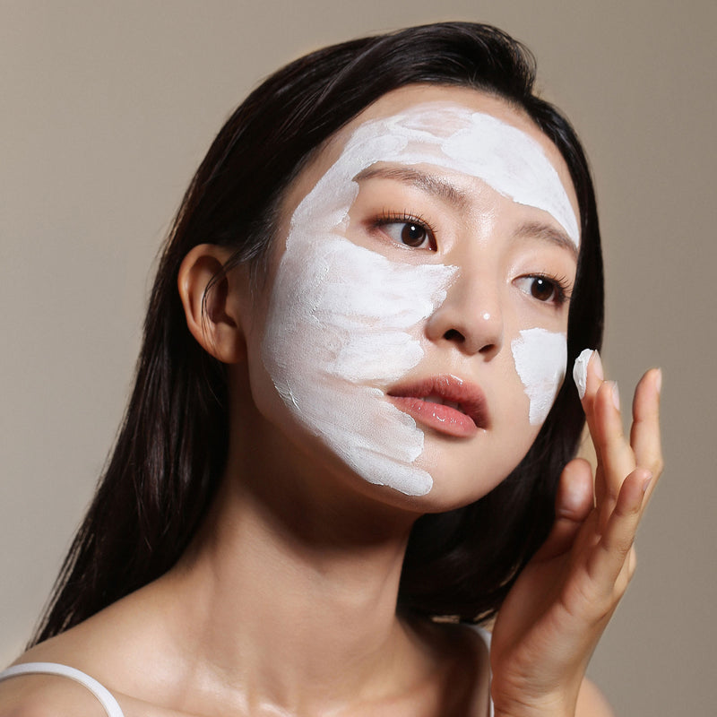 Asian woman with white cream spread onto forehead, cheek, and half of her chin as she uses her finger to spread it onto her other cheek in front of a light brown background.