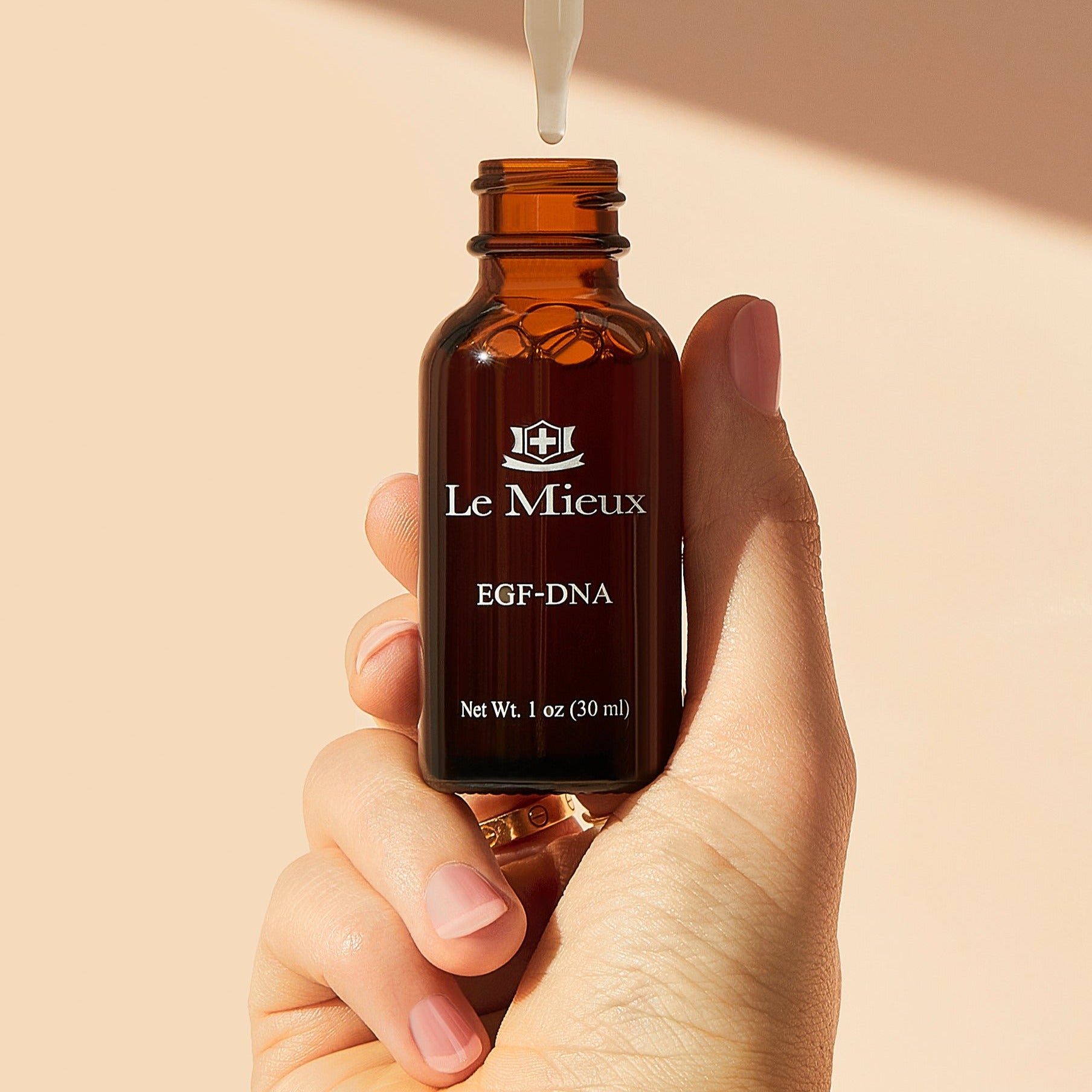 hand holding an uncapped amber EGF-DNA bottle with the dropper hovering above it in front of a light peach background