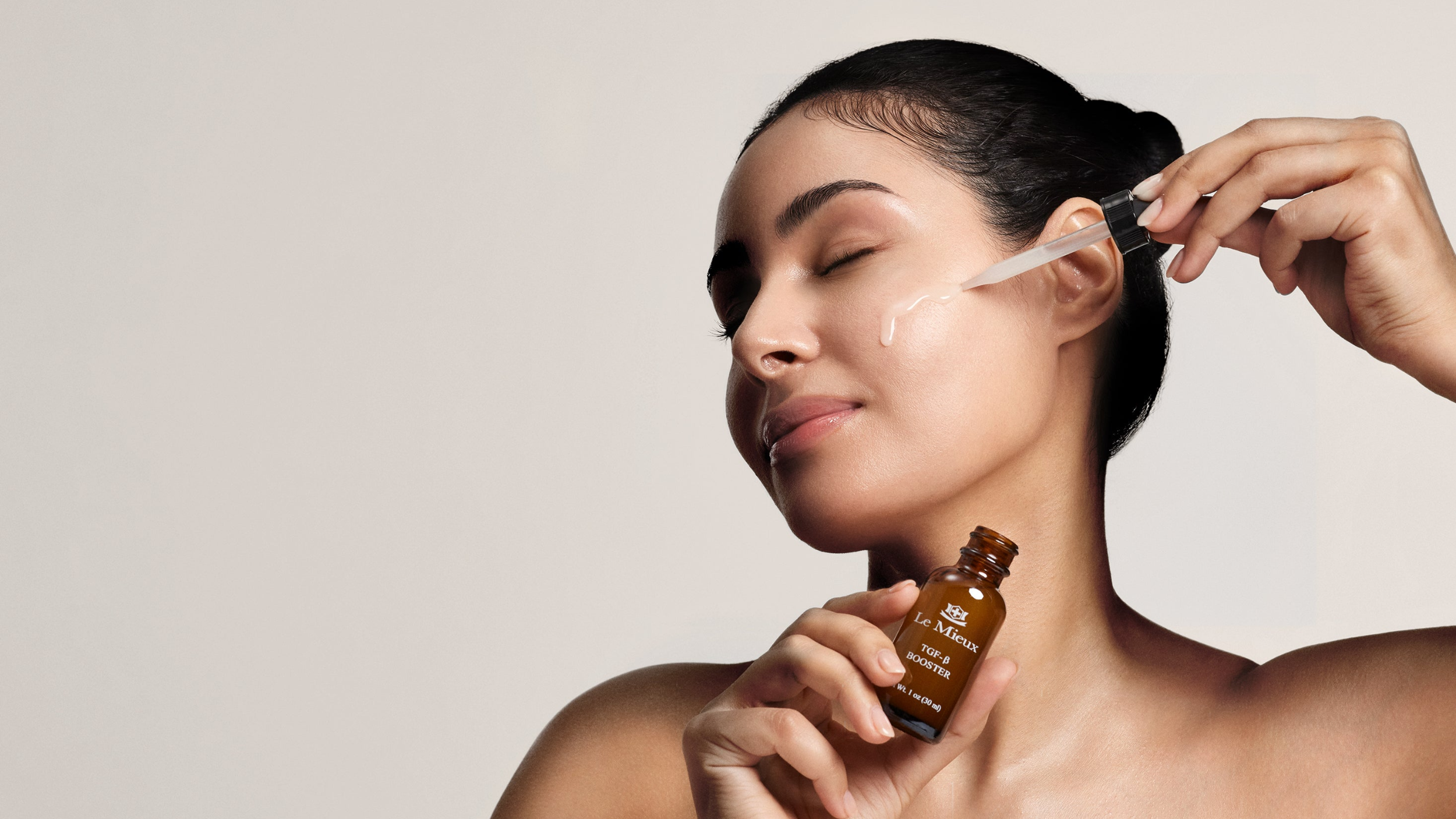 Woman applying a dropper of TGF-β Booster serum on face against a neutral background