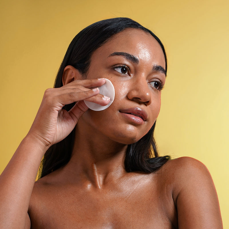 Woman in front of a yellow background with damp white cotton pad pressed to her cheek.